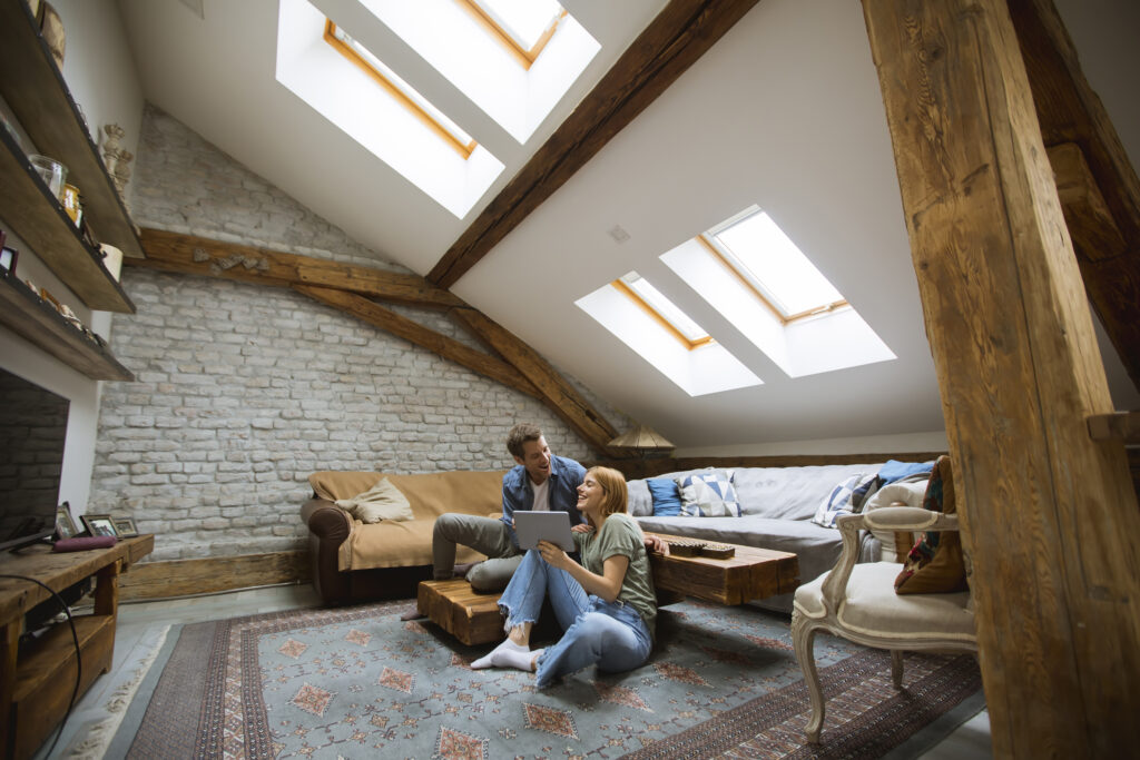 ein junges paar sitzt unter 4 großen hellen dachfenstern in einem wohnzimmer im dachgeschoss.